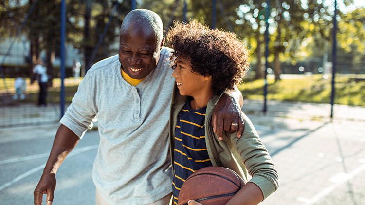 一个男孩拿着一个篮球，和他的祖父手挽着手走过篮球场.