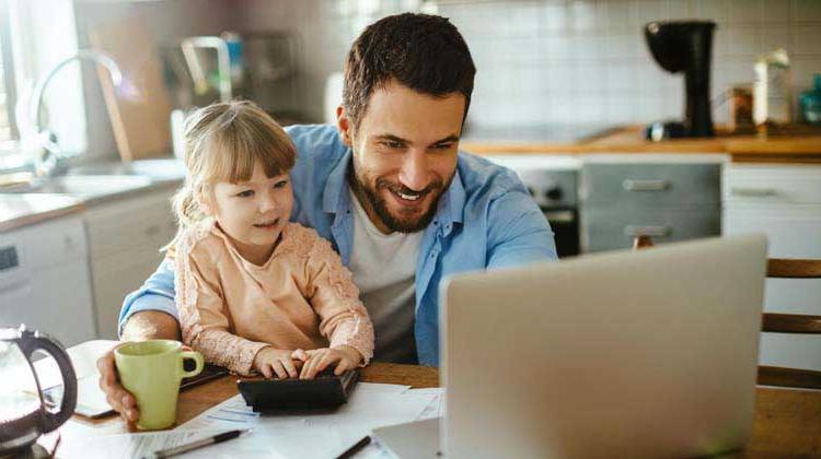 一个男人和他的女儿坐在他的腿上，坐在厨房的桌子上，用笔记本电脑研究存钱的理由和不同种类的储蓄账户.